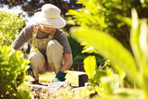 A woman wearing a wide-brimmed hat, beige trousers, and gardening gloves is kneeling on a neatly maintained lawn in a garden, actively planting or tending to the soil near a flower bed border. The garden features a lush, green grassy area with dense foliage and shrubbery in the background, including trees and flowering plants, indicating a landscaped outdoor space. The bed is edged with soil and possibly stone or brick borders, and there are garden tools, such as a trowel, visible on the ground. The scene is illuminated by natural sunlight, suggesting a bright, clear day with warm weather, typical of outdoor gardening in St John's Wood. This image exemplifies professional gardening services provided by Lawn Mowing St Johns Wood, highlighting aspects of lawn care, planting, and garden maintenance in a well-kept, suburban garden environment.