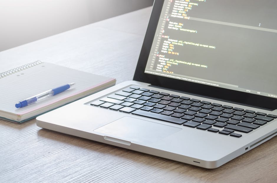 A silver laptop displaying lines of code on a dark background sits open on a light wooden desk, next to an open spiral notebook with a blue pen resting on it. The scene is illuminated by natural light
